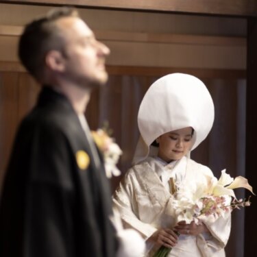 手前の新郎をぼかし、綿帽子の白無垢の花嫁がブーケを見つめる姿を能舞台で写した和装前撮り