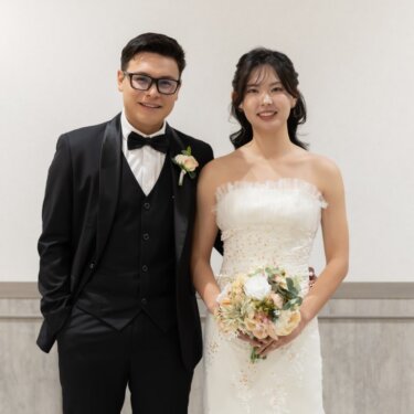 壁灯のある白い壁の前に並んで立ち、ほほえむタキシードの新郎とウェディングドレスの花嫁の正面の結婚写真