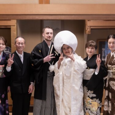 和室で綿帽子の白無垢の花嫁と新郎を囲み、家族全員がピースサインで笑う家族写真の和装前撮り