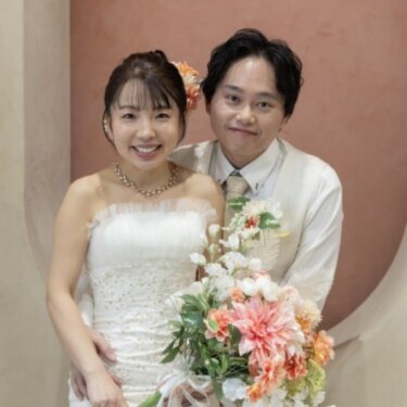 テラコッタ色の壁の前で、ピンクのダリアのブーケを持つ花嫁と後ろから寄り添う新郎のスタジオ前撮り