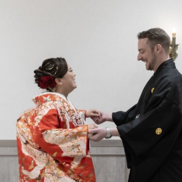 壁灯のある白い部屋で手を取り合い、大笑いする鶴柄の赤い色打掛の花嫁と紋付袴の新郎。ファーストミートの和装前撮り