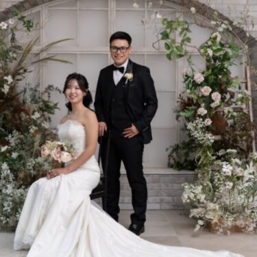 レンガ造りのアーチ窓と花々のアレンジを望むガーデンスタジオで、椅子に座る花嫁の長いトレーンが美しいフォトウェディングの一枚