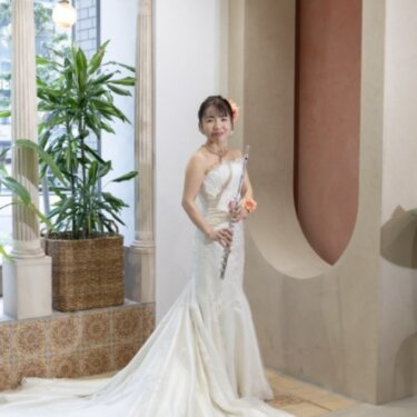 アーチ窓と観葉植物のそばで、フルートを手に立つマーメイドラインのウェディングドレスの花嫁。趣味の楽器と撮る前撮り