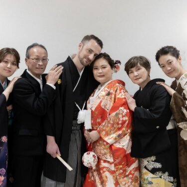 壁灯のある白い部屋で、和装の家族全員と寄り添う新郎新婦。前撮りで残す賑やかな家族写真