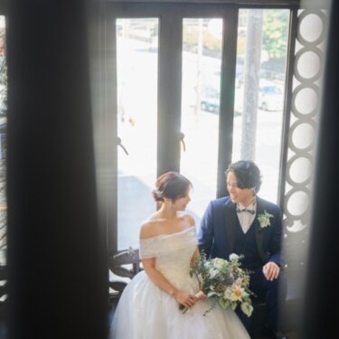 カーテン越しに、大きな窓辺に並んで座り見つめ合う新郎新婦を写したのぞき見構図のウェディングフォト