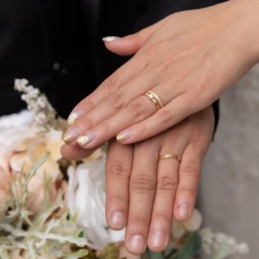 ブーケの上で重ねた二人の手に光るゴールドの結婚指輪のクローズアップ。前撮りのディテールカット