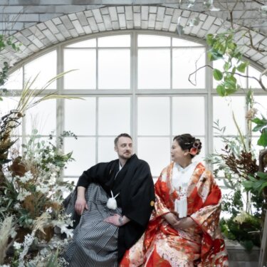 レンガ造りのアーチ窓と花々のガーデンスタジオで、座って見つめ合う新郎新婦を引きで写したスタジオフォト