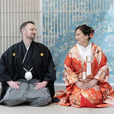 青い花柄の壁と格子のセットで正座し、見つめ合う紋付袴の新郎と赤い色打掛の花嫁の和装フォトウェディング