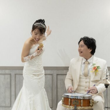ドラムを叩く新郎を見て大笑いするマーメイドドレスの花嫁。趣味の演奏を取り入れた結婚写真
