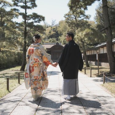 松並木の石畳の参道を手をつないで歩く、色打掛の花嫁と羽織袴の新郎の後ろ姿。三渓園で撮る和装フォトウェディング