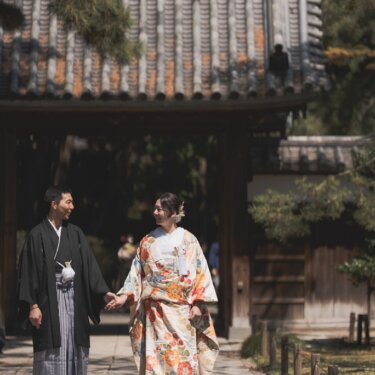 三渓園の山門をくぐり、手をつないでこちらへ歩いてくる新郎新婦。松の緑と色打掛が映える和装前撮り