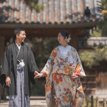 瓦屋根の門の前で手をつなぎ、大きく笑って見つめ合う黒紋付袴の新郎と花柄の色打掛の花嫁の結婚写真