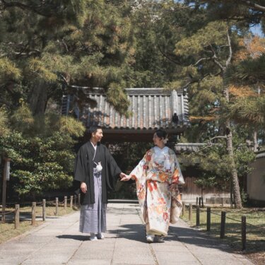 三渓園の松並木の参道を、瓦屋根の門を背景に手をつないで歩く新郎新婦の和装ロケーションフォト