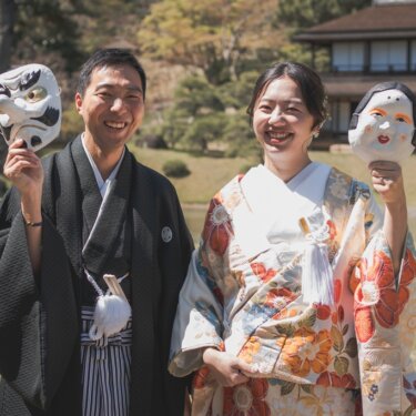 天狗とおかめのお面を掲げて笑う新郎新婦。芝生と日本家屋を背景にした遊び心あるお面の和装フォトウェディング