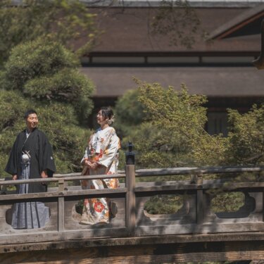 三渓園の石の欄干の橋で離れて立ち、笑い合う黒紋付袴の新郎と色打掛の花嫁。趣ある日本家屋を背景にした和装ロケーションフォト