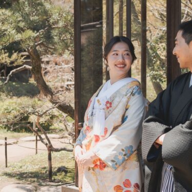 格子の窓辺で肩を寄せ、見つめ合って笑う花柄の色打掛の花嫁と黒紋付の新郎。庭の木々を背景にした結婚写真