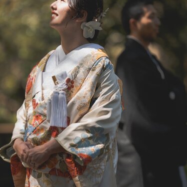 空を見上げて笑う色打掛の花嫁と、後ろに立つ新郎。白い花の髪飾りが印象的な和装前撮り