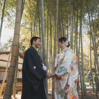 三渓園の竹林で茅葺屋根の建物を背に、両手を取り合って見つめ合う黒紋付袴の新郎と花柄の色打掛の花嫁の和装前撮り