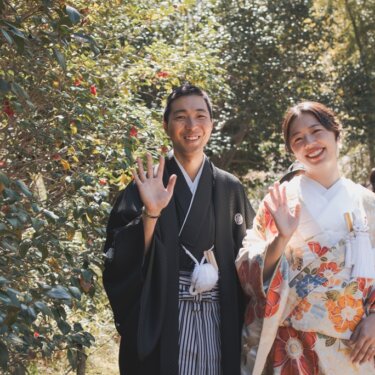 三溪園にて、赤い椿の咲く木々を背景に、手を振ってカメラに笑顔を向ける羽織袴の新郎と色打掛の花嫁のフォトウェディング