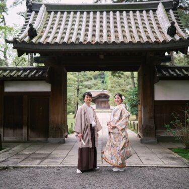 三渓園の瓦屋根の山門をくぐった先で、カメラに並んで笑う羽織袴の新郎と色打掛の花嫁の前撮り