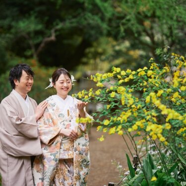 黄色い花が咲く木立を眺めて笑う新郎新婦。新郎が花嫁の肩に手を添える春の和装フォトウェディング