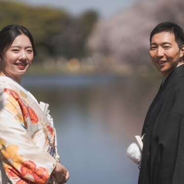 三渓園の池のほとりで振り返って笑う新郎新婦。桜と水面を背景にした色打掛の結婚写真