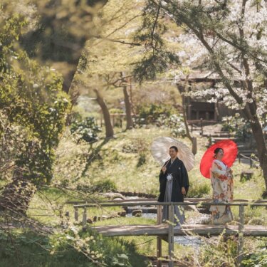 桜の木々に囲まれた小さな橋の上で、白と朱赤の和傘を差す新郎新婦を引きで写した和装前撮り