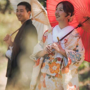 朱赤の和傘を差して空を見上げ笑う色打掛の花嫁を、新郎の白い和傘越しに写した春の和装前撮り