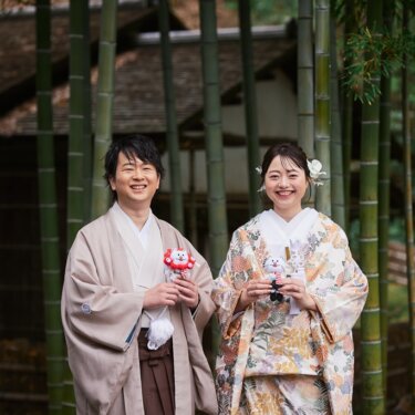 竹林と茅葺屋根の建物の前で、小さなぬいぐるみを手にカメラへ笑顔を向ける色打掛の花嫁と羽織袴の新郎の前撮り