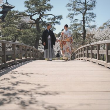 三渓園の旧燈明寺三重塔を望む橋を手をつないで歩く新郎新婦。満開の桜が咲く春の和装前撮り