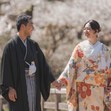 桜をぼかした橋の上で、手をつなぎ見つめ合って笑う羽織袴の新郎と色打掛の花嫁の結婚写真
