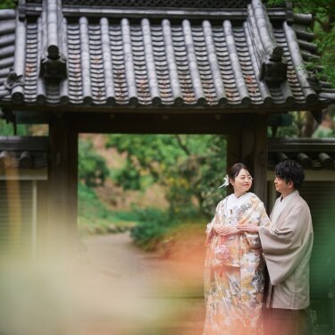 瓦屋根の門を背景に、前景の草花をぼかして写した寄り添う新郎新婦。木漏れ日が差す三渓園の和装ロケーションフォト
