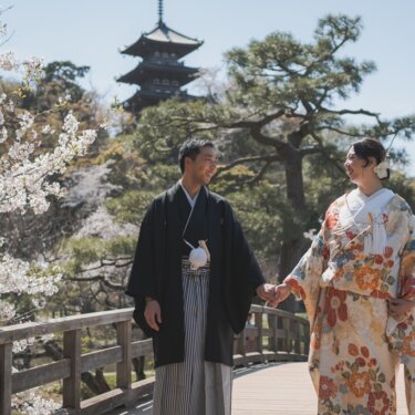 三重塔を望む木の反り橋で手をつなぎ見つめ合う新郎新婦。桜の枝が手前に写る三渓園の春のロケーションフォト