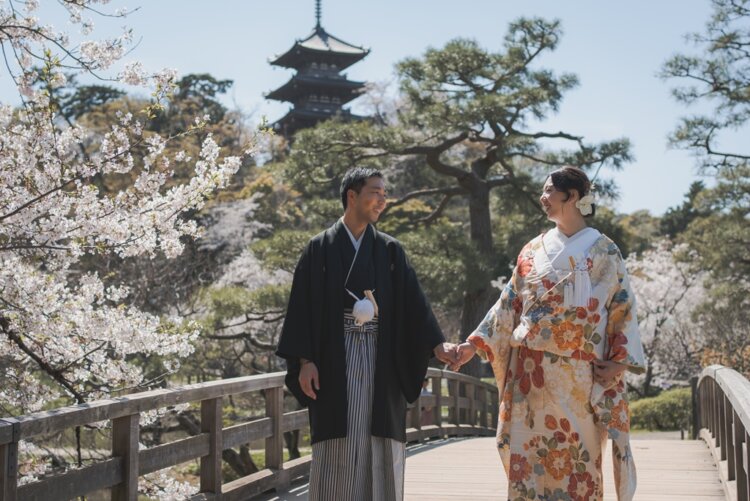 三重塔を望む木の反り橋で手をつなぎ見つめ合う新郎新婦。桜の枝が手前に写る三溪園の春のロケーションフォト