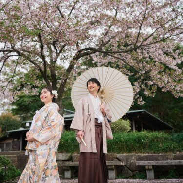 満開の桜を背景に、白い和傘を持つ羽織袴の新郎と笑う色打掛の花嫁。東屋の屋根がのぞく三渓園の和装前撮り