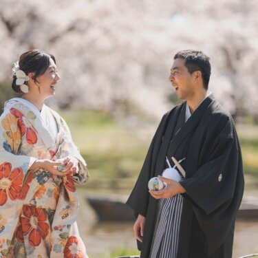 池の小舟と満開の桜を背景に、手毬を手に見つめ合う新郎新婦。春の色打掛で撮る和装ロケーションフォト