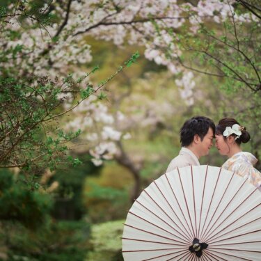 白い和傘の陰で額を寄せ合う新郎新婦。桜の枝に包まれた庭園で撮るロマンチックな前撮り