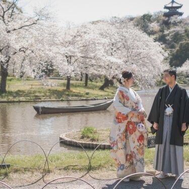 満開の桜と池に浮かぶ小舟、遠くの三重塔を望む三渓園で、向かい合う色打掛の花嫁と羽織袴の新郎を引きで写した和装前撮り