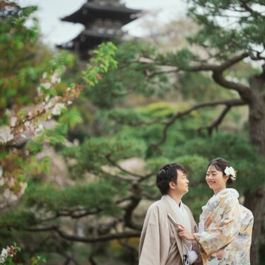 三渓園の三重塔を見上げる橋の欄干で、寄り添って笑う色打掛の花嫁と羽織袴の新郎。松と桜に囲まれた春の和装前撮り