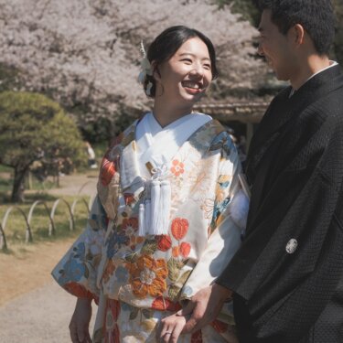 桜咲く木々を背景に手を握り合い、見つめ合って笑う色打掛の花嫁と黒紋付の新郎の結婚写真