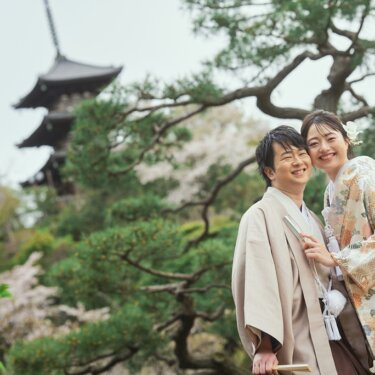 三重塔と松を背景に肩を寄せ合い、カメラに笑顔を向ける色打掛の花嫁と羽織袴の新郎の結婚写真