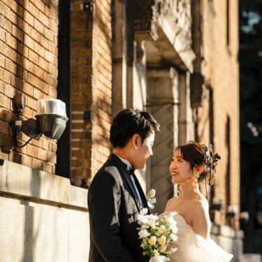 歴史的なレンガ建築の壁の前で、白いブーケを持って見つめ合う新郎新婦。オフショルダーのドレスで撮る前撮り