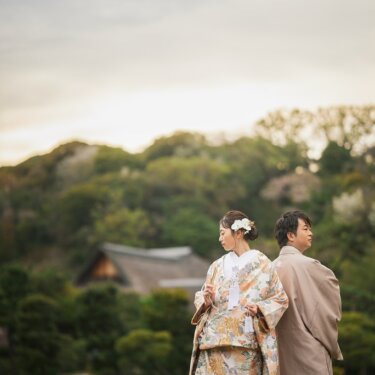 茅葺屋根の建物を遠くに望む池のほとりで背中合わせに立つ、色打掛の花嫁と袴姿の新郎の和装ロケーションフォト
