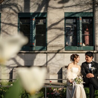 白いチューリップを前景に、花壇の柵に座って見つめ合う新郎新婦。石造りの洋館の街並みが華やかな前撮り