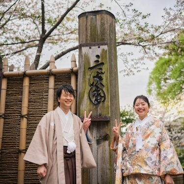 '三渓園'の木柱と竹垣の前でピースサインをする、ピンクベージュの色打掛の花嫁と羽織袴の新郎。桜の季節の和装前撮り
