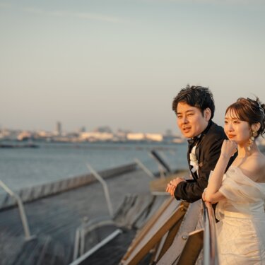 大さん橋の柵際で遠くの海と横浜港を眺める、タキシードの新郎とウェディングドレスの花嫁の結婚写真
