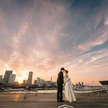 大さん橋のウッドデッキで、夕焼けに染まるみなとみらいのビル群を背にキスをする新郎新婦を引きで写した前撮り