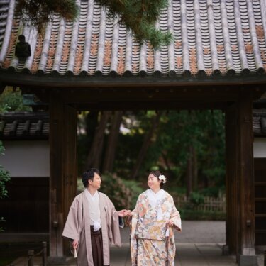 三渓園の山門の下で手をつなぎ、見つめ合って笑う新郎新婦。石畳と門が格式を感じさせる和装前撮り
