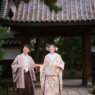 瓦屋根の門の前で手をつなぎ、大きく笑って空を見上げる羽織袴の新郎と色打掛の花嫁の結婚写真