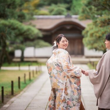 松並木の参道で手をつなぎ、振り返ってカメラに笑顔を向ける色打掛の花嫁。背中の刺繍が美しい和装前撮り